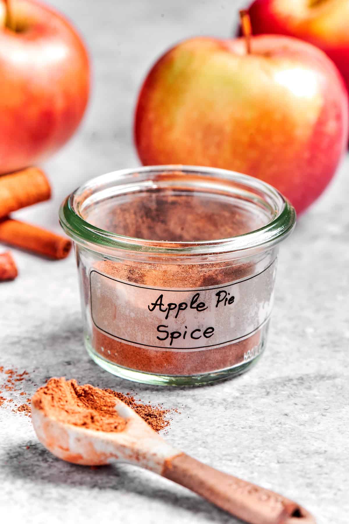 Apple Pie Spice brings some fall magic to all kinds of recipes and my version is a slightly different blend with extra warmth and coziness! Jar of apple pie spice with spoonful of spice in foreground and apples in background