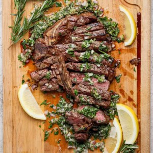 Overhead view of sliced grilled t-bone steak with board sauce, fresh rosemary, and lemon wedges
