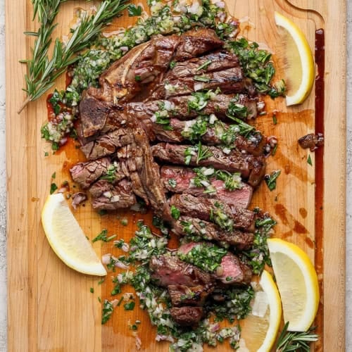 Overhead view of sliced grilled t-bone steak with board sauce, fresh rosemary, and lemon wedges