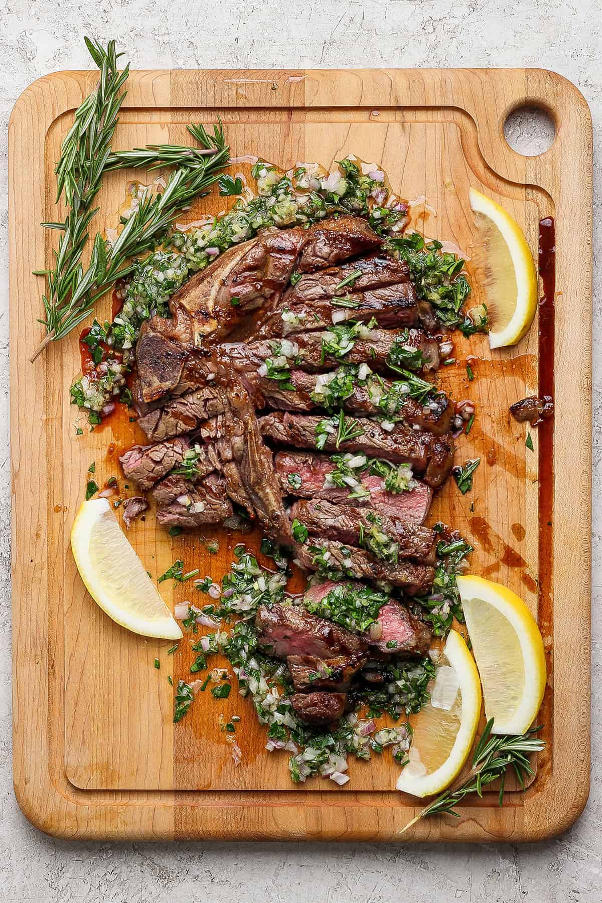 Overhead view of sliced grilled t-bone steak with board sauce, fresh rosemary, and lemon wedges