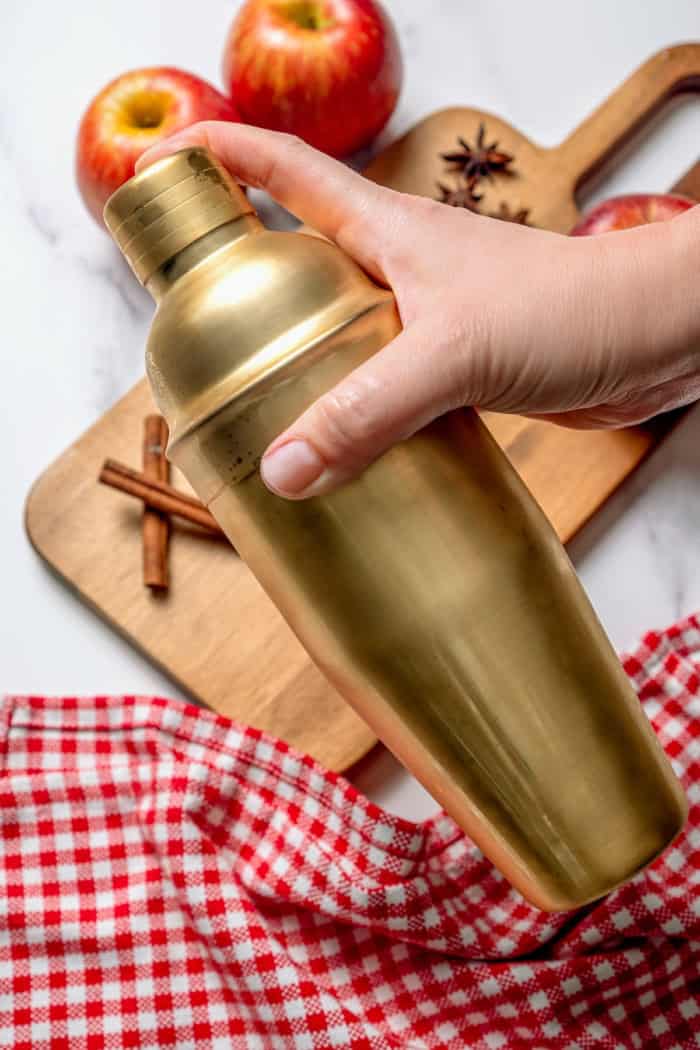 A fall spin on the classic, this Apple Cider Margarita is lightly sweetened with maple syrup and a cinnamon rim add a festive touch! Hand shaking apple cider margarita in gold cocktail shaker.