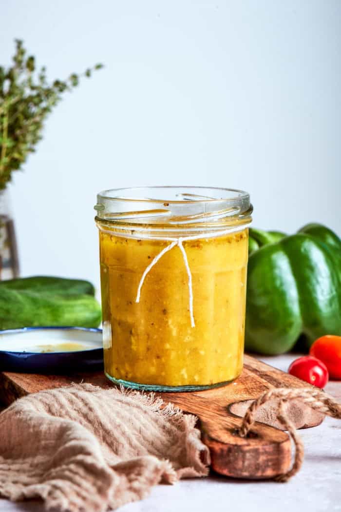 Bright and tangy, with lots of garlic and oregano, this Greek Salad Dressing is easy to make and tastes SO much better than store-bought! Jar of Greek salad dressing on wood board with vegetables in background