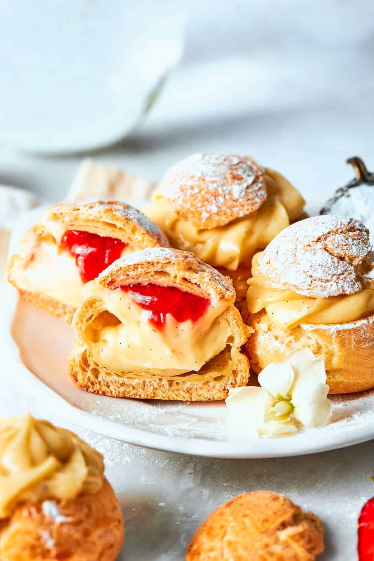 mini cream puffs filled with vanilla pastry cream on a white plate, one is cut in half to reveal strawberry sauce