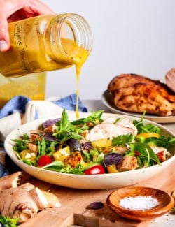 a hand pouring ginger dressing, from a jar, over a fresh green salad with grilled chicken
