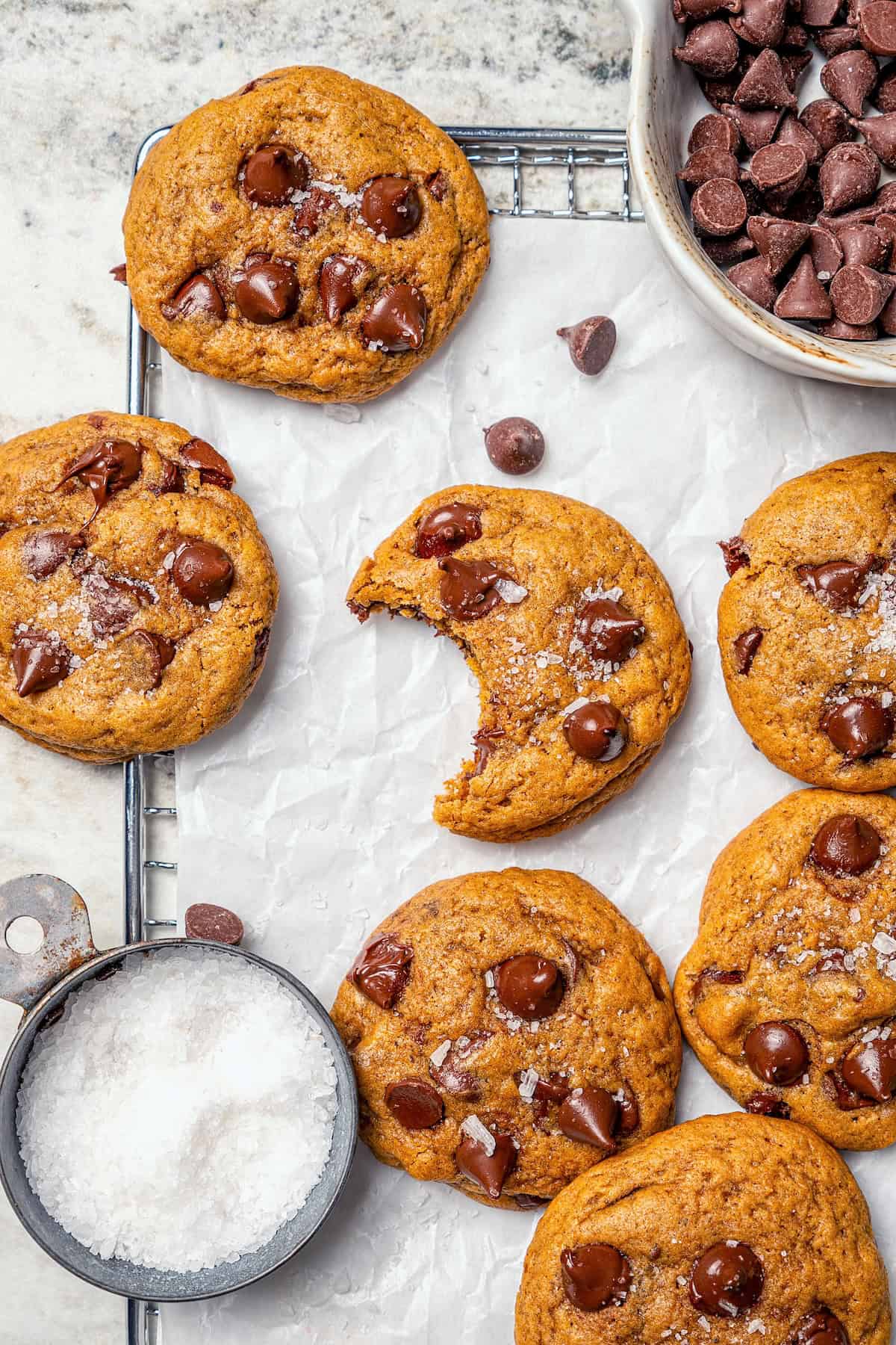 Soft, moist, and chewy, these Pumpkin Chocolate Chip Cookies are the perfect sweet treat for fall! Pumpkin pie spice adds warm, cozy flavor. Pumpkin chocolate chip cookies on parchment lined wire rack with bowls of chocolate chips and sea salt.