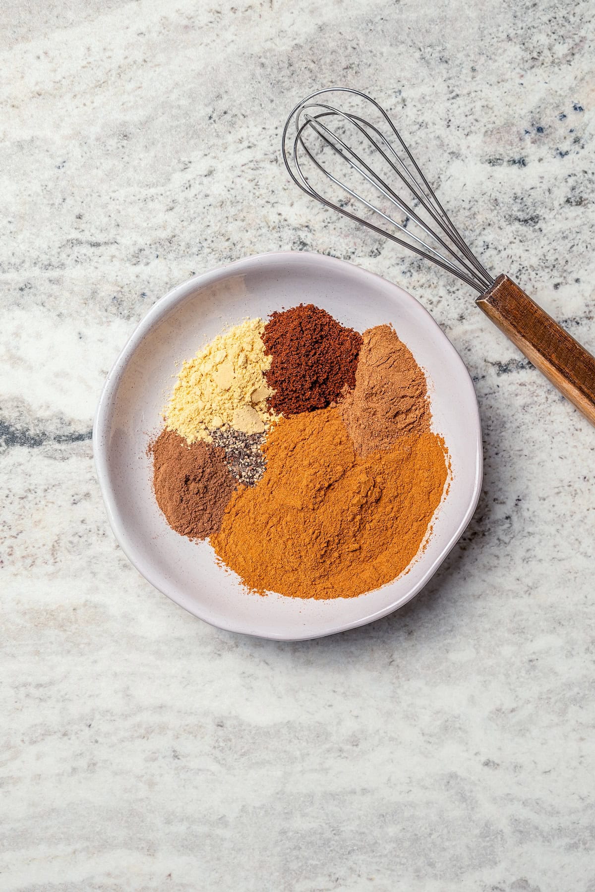This isn't your ordinary homemade Pumpkin Pie Spice recipe! Itβs rich and robust, with a touch of black pepper for some cozy warmth. Overhead view of spices for homemade pumpkin pie spice on plate with whisk.