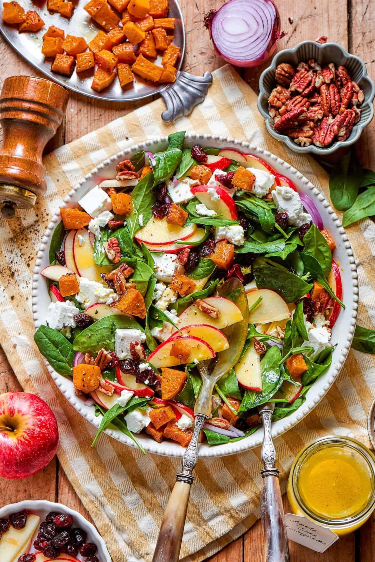 This easy harvest salad combines roasted butternut squash with maple pecans, cranberries, and goat cheese, tossed in a curry fig vinaigrette. overhead photo of a wood table with a platter full of fall harvest salad with fresh greens, butternut squash, fresh greens, pecans, cranberries, and goat cheese