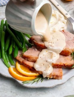 a hand pouring a gravy pitcher of ham gravy over slices of ham on a plate
