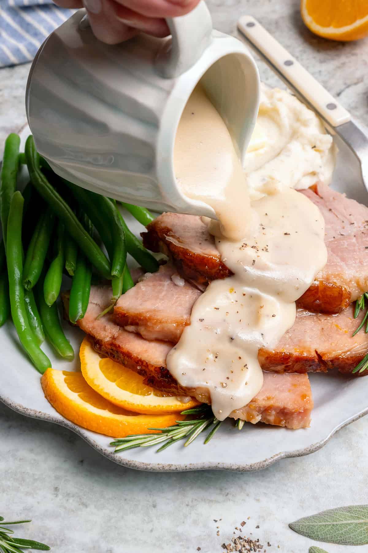 a hand pouring a gravy pitcher of ham gravy over slices of ham on a plate