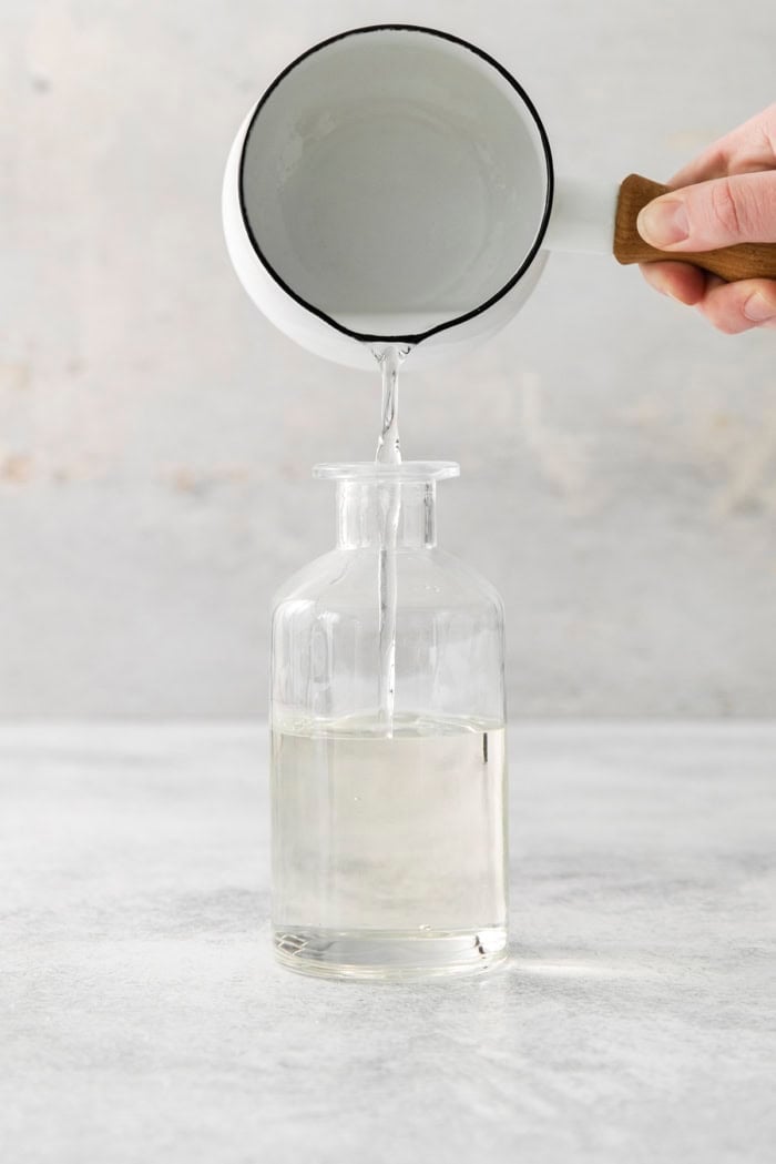 a hand pouring simple syrup from a small white pan into a clear bottle