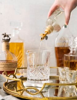 a hand pouring simple syrup from a clear bottle into a cocktail mixing glass