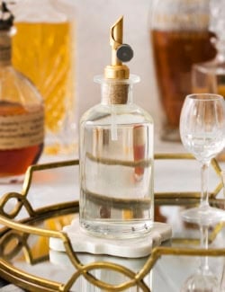 a clear bottle with a gold pour spout, full of simple syrup, sitting on a gold mirrored cocktail tray