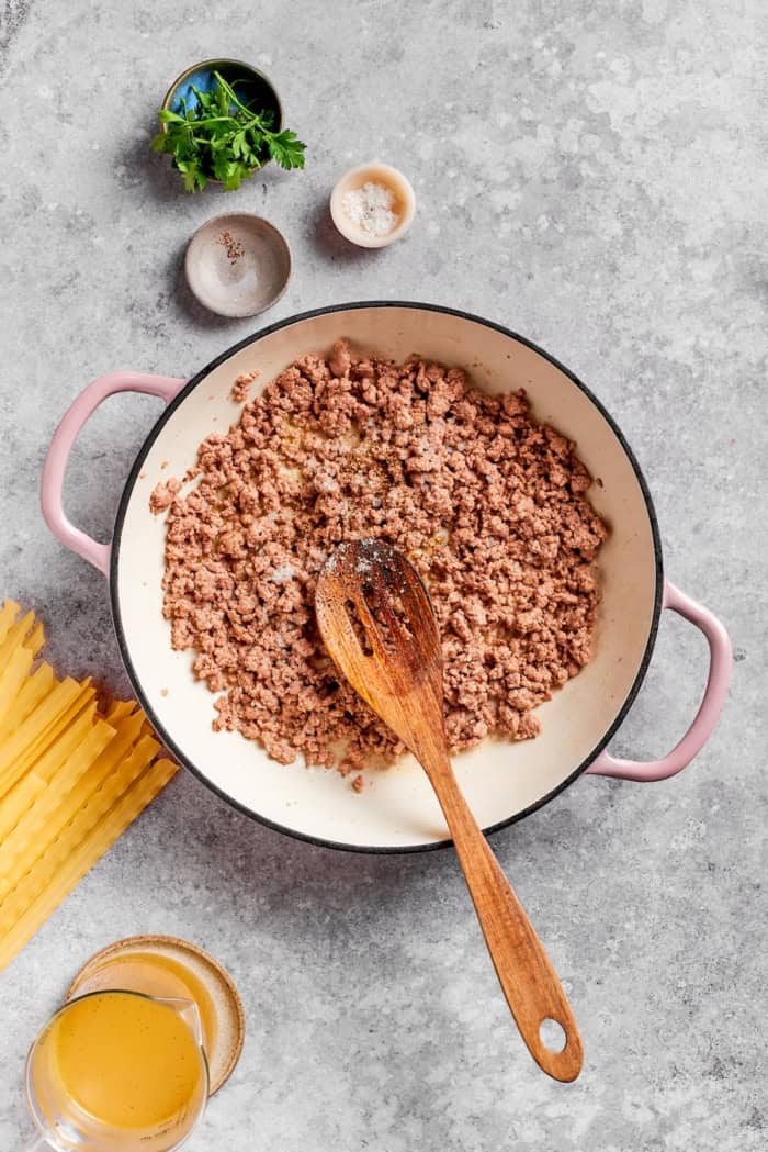 This creamy Ground Beef Stroganoff recipe is made from scratch without canned soup! An easy, budget-friendly dinner your family will love. Ground beef in skillet with wooden spoon.