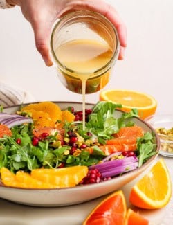 a hand pouring citrus vinaigrette over a bowl of salad greens with fresh citrus slices