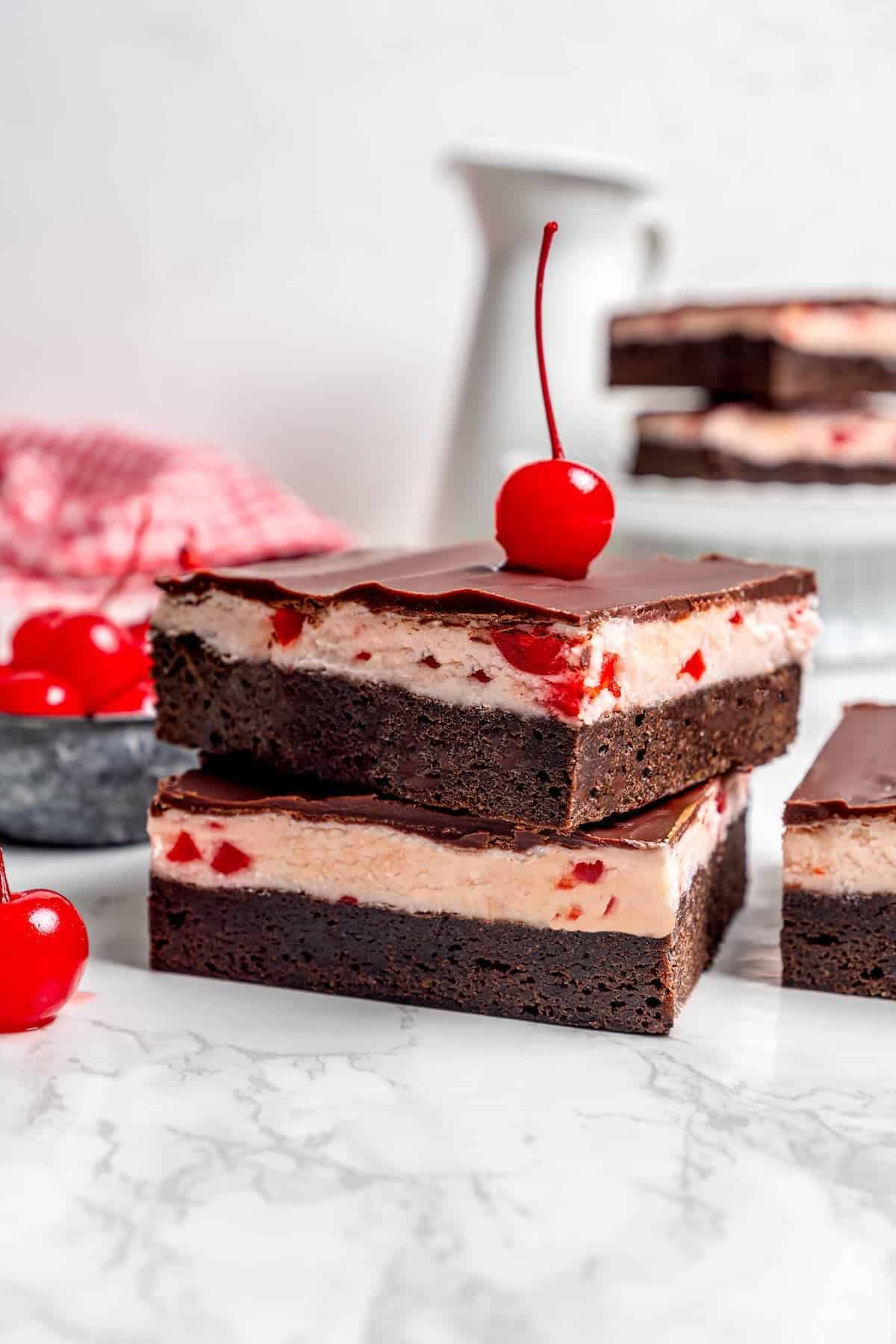 These fudgy cherry brownies are layered with fluffy cherry buttercream and smooth chocolate glaze for an indulgent bakery-style dessert! side view of a stack of cherry chocolate brownies with a maraschino cherry on top
