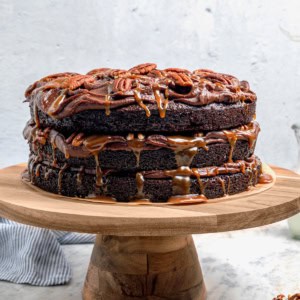 a layered chocolate, caramel, and pecans Turtle Cake on a wooden cake pedestal