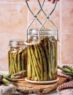 a hand setting a jar of pickled asparagus down onto a wood cutting board, using a jar lifter tool