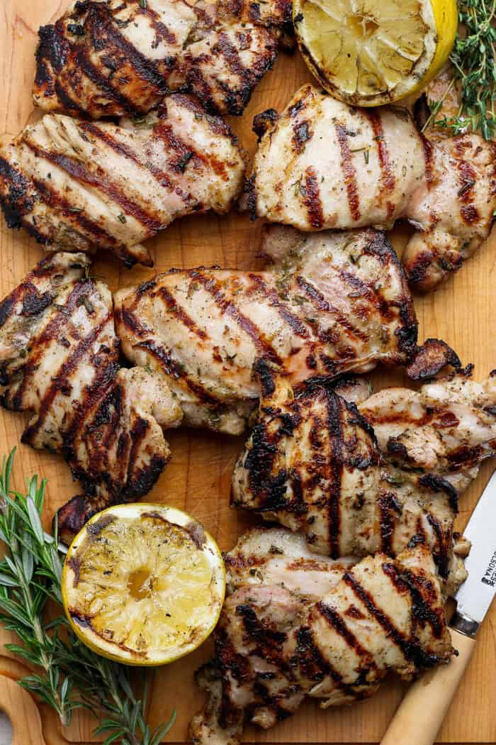 These grilled lemon chicken thighs are juicy and smoky, with a fresh lemon-herb marinade to brighten things up. The perfect summer main dish! Grilled lemon chicken thighs on cutting board with fresh herbs and charred lemon halves.