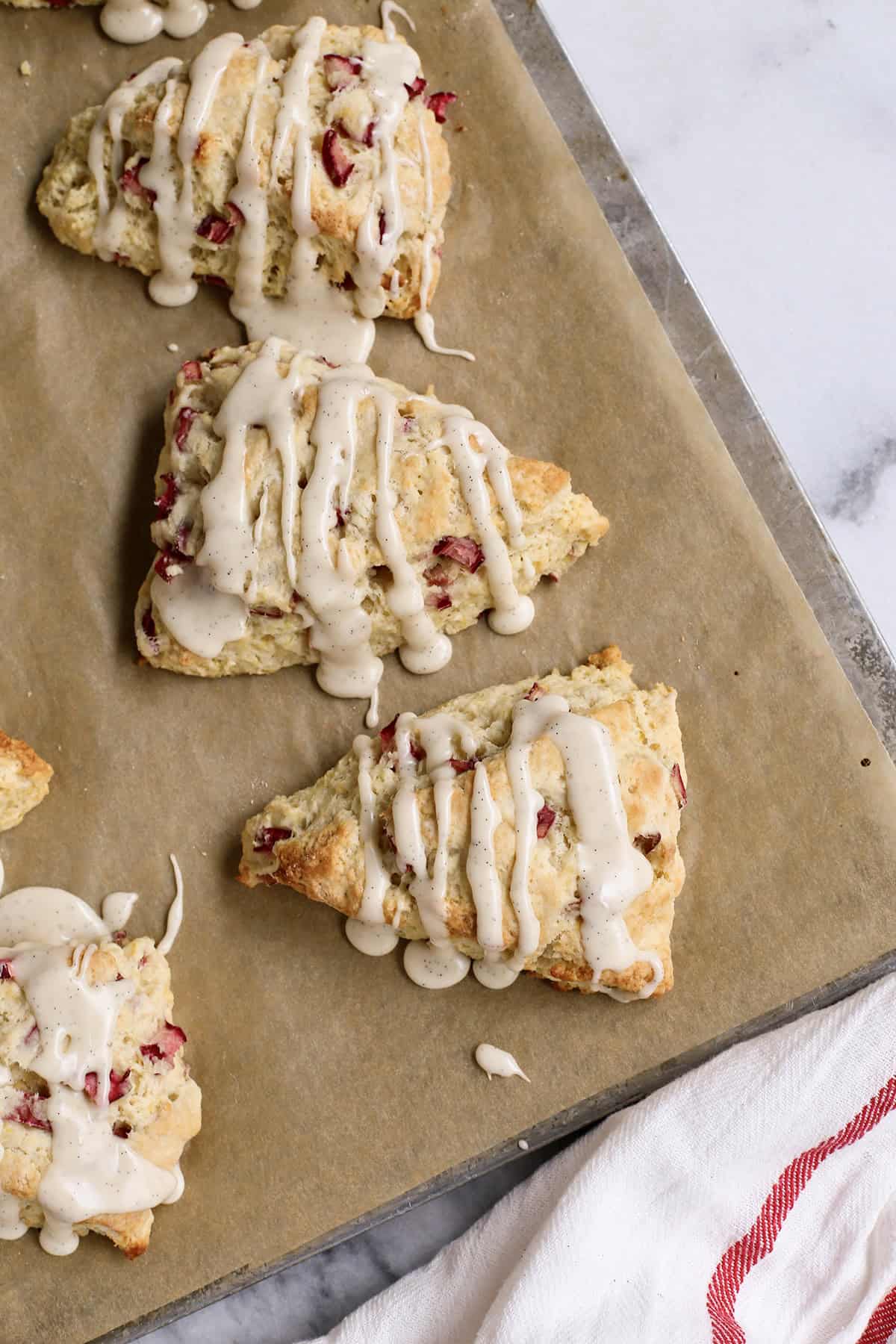 These rhubarb scones are moist inside and flaky outside, filled with juicy tart rhubarb and fresh lemon zest. Perfect with coffee or tea! overhead photo of baked rhubarb scones with glaze, on a baking sheet with parchment paper