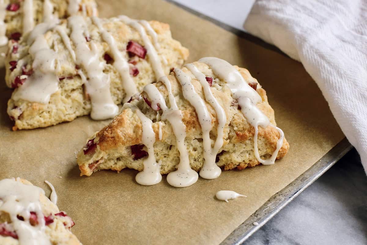 These rhubarb scones are moist inside and flaky outside, filled with juicy tart rhubarb and fresh lemon zest. Perfect with coffee or tea! baked rhubarb scones with glaze, on a baking sheet with parchment paper