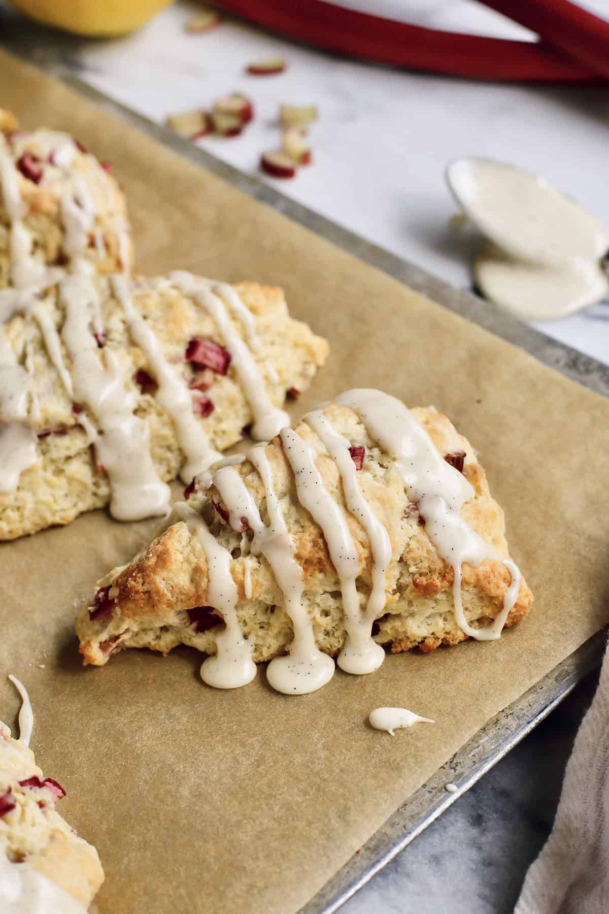 These rhubarb scones are moist inside and flaky outside, filled with juicy tart rhubarb and fresh lemon zest. Perfect with coffee or tea! baked rhubarb scones with glaze, on a baking sheet with parchment paper