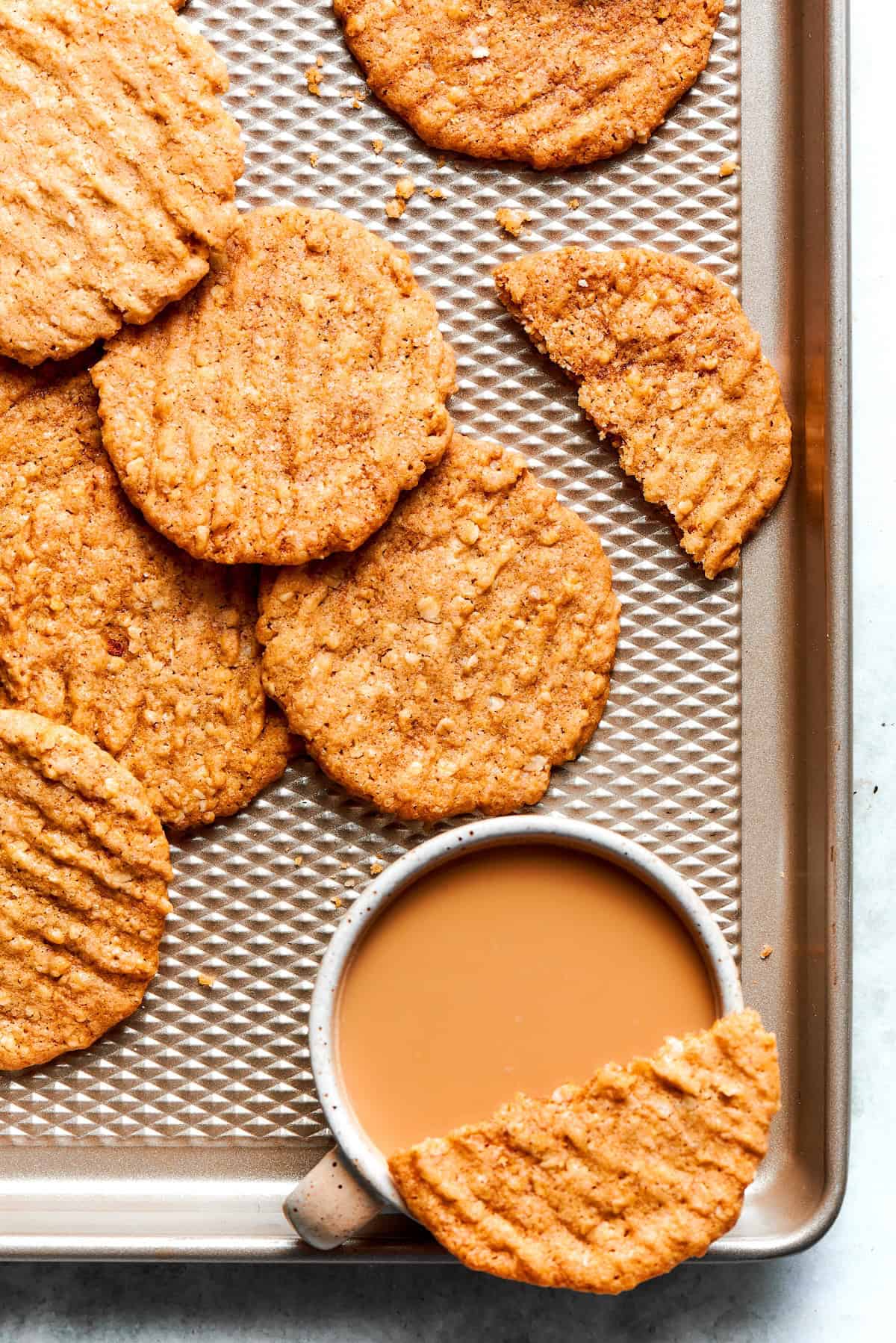 These thin, crispy oat cookies are buttery and golden on the edges, with the most delightful crunch. Made with simple pantry ingredients! Crispy oat cookies on sheet pan with cup of coffee.